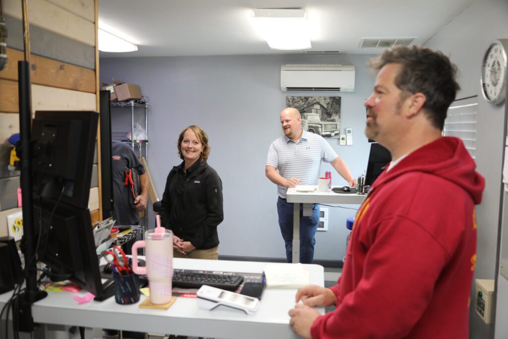 Employees interact cheerfully in a clean, organized office area of an auto repair shop. With computer stations, paperwork, and tools visible, the scene reflects a welcoming and professional customer service environment.