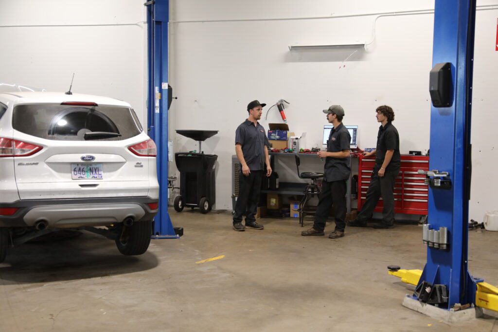 Three auto technicians stand and converse near a red tool chest and computer workstation inside a clean, well-lit garage. A white Ford Escape with Oregon plates is parked nearby under a hydraulic lift.