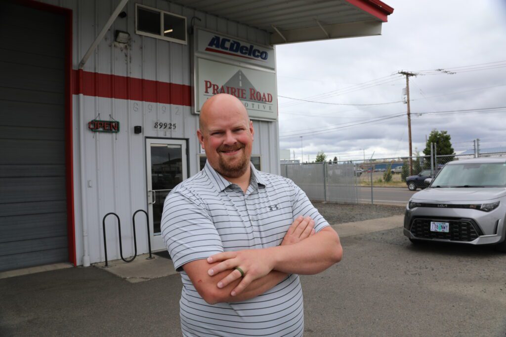 A smiling man stands confidently with arms crossed outside Prairie Road Automotive, next to the shop’s entrance under ACDelco signage. The clean exterior and welcoming posture convey professionalism and customer-friendly service.